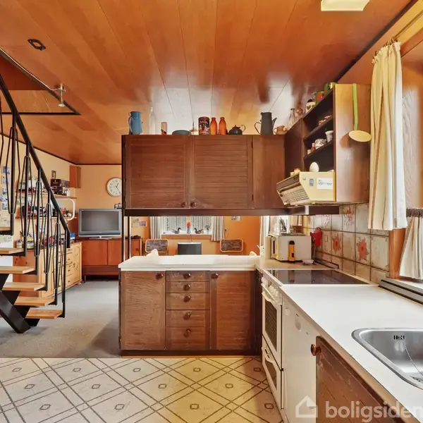 Et køkken med et hvidt komfur og vask er omgivet af træpaneler. En trappe fører op, og der er et vindue med udsigt og blomster i karmen. Skabsplads øverst.