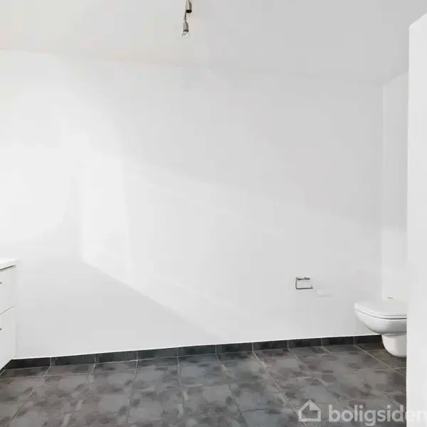 En rummelig badeværelse med mørke flisegulve. Hvidt skabsarrangement til venstre og vægmonteret toilet til højre. Rummet er lys med en minimalistisk indretning.