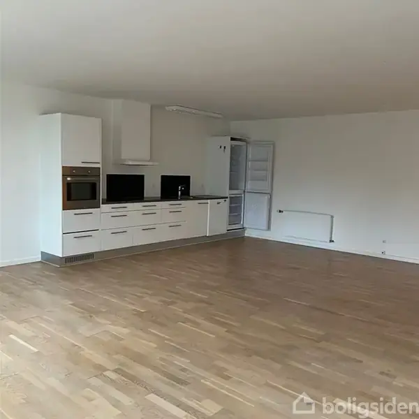 Et moderne køkken med hvide skabe og indbygget ovn står langs en væg. Køleskabet er åbent. Rummet har trægulv og er lysfyldt med hvide vægge.