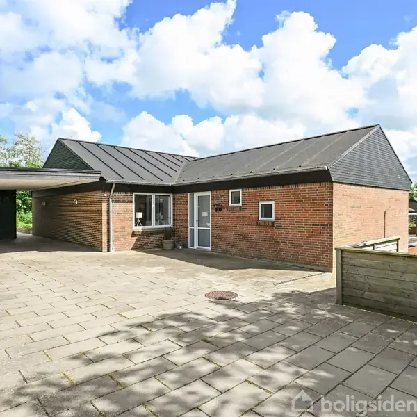 En enkeltliggende, murstenshus med carport ved siden af, står på en flisebelagt indkørsel. Huset har sorte tagplader og et velplejet hegn omkranser ejendommen.