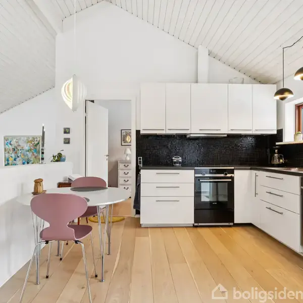 Et moderne køkken med hvide skabe og sort backsplash. Et lille rundt bord med to lyserøde stole står i forgrunden. Rummet har lyse trægulve og hængelamper.