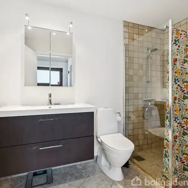 Et badeværelse med et toilet og en vask med spejlskab. Bruseren er til højre og adskilles af et farverigt blomsterforhæng. Rummet har lys beige fliser på væggen.