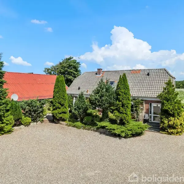 Et rødt tegltagshus omgivet af høje grønne buske og træer står på en grusbelagt gårdsplads. Huset har et vindue og en dør mod gårdspladsen.