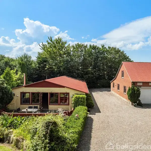 Huset med rødtag ligger ved siden af en bred grusvej omgivet af frodigt grøntområde og træer under en blå himmel med skyer.
