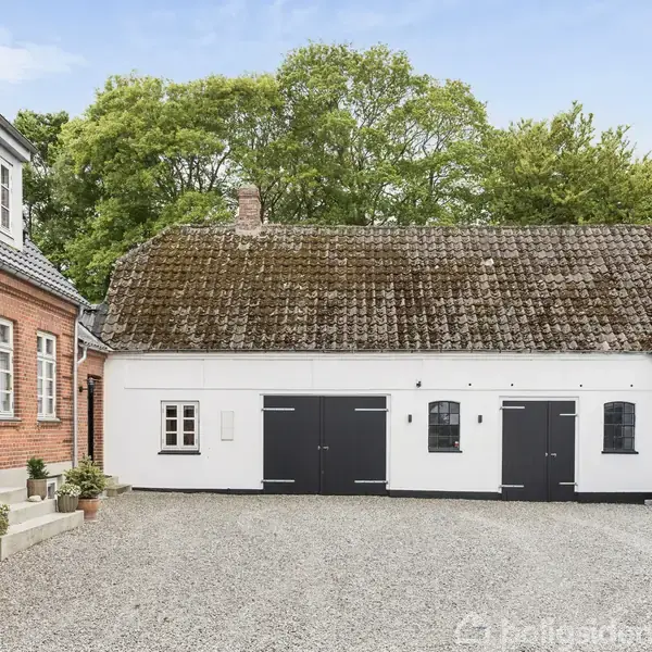 Et gårdhus med røde mursten og hvid facade, der står omkring en gårdsplads med grus. Sorte døre og vinduer pryder bygningerne. Træer ses i baggrunden.