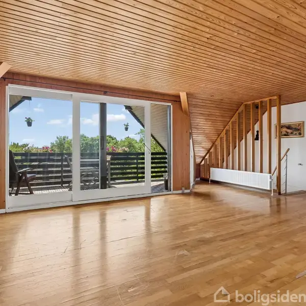 Trægulv dominerer et rum med skrånende træloft og store glasdøre, der åbner ud mod en balkon med udsigt over træer. Trappen fører til en overetage.