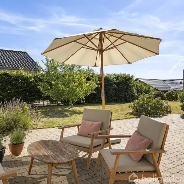 Terrasse med to loungestole under en stor parasol. Et rundt træbord står mellem stolene, omgivet af planter i potter. I baggrunden er der et spisebord og have med hække.