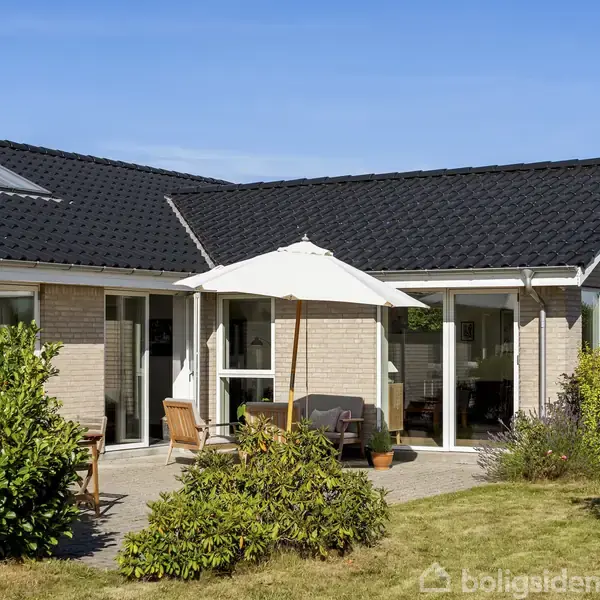Et moderne hus med lyse mursten står i sollys, flankeret af en grøn have. En parasol og havemøbler er placeret på terrassen foran huset, omgivet af buske.