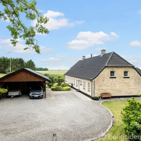 Et stort, hvidt hus med sort tag står omkranset af græsarealer. Til venstre ses en carport med en bil parkeret. Landskabet består af åbne marker og træer i baggrunden.