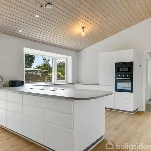 Et moderne køkken med hvidt skabsinventar og en stor køkkenø. Der er indbyggede ovne og et bredt vindue, der giver udsigt til en grøn have.