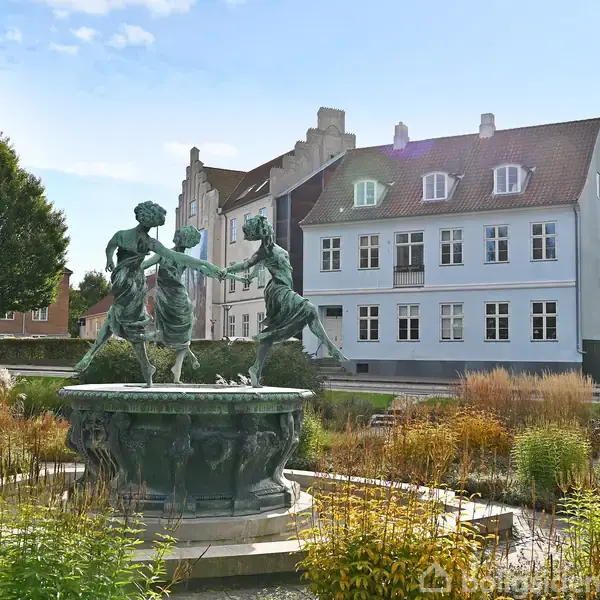 En bronze skulptur af to dansende figurer oven på en springvand. Omkranset af beplantning i en park med en historisk bygning i baggrunden.