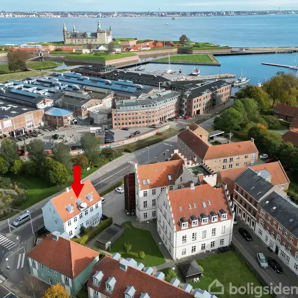 En hvid bygning med rødt tag markeres med en rød pil; den ligger ved en vej omgivet af andre bygninger. I baggrunden ses en havn med et historisk slot ved kysten.