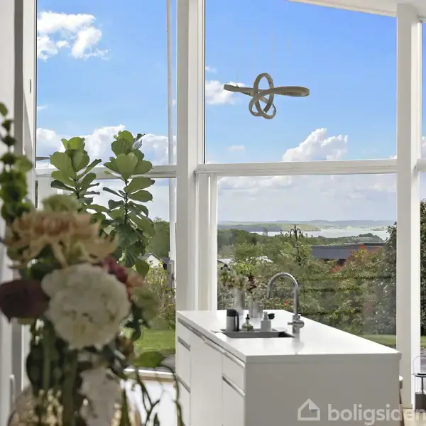 Køkkenbord med vask står foran store vinduer, der afslører en udsigt til en have og en fjord under en blå himmel. Blomster dekorerer forgrunden.