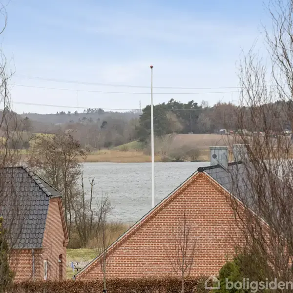 To tage af røde murstenshuse med flagstang ses i forgrund, omgivet af trægrene. I baggrunden ligger en sø og en skovklædt bakkeskråning under en overskyet himmel.