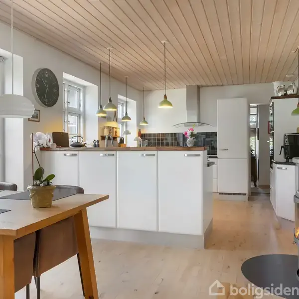 Moderne køkken med hvidt skabsmøblement og brændeovn. Langt spisebord med stole står i forgrunden. Loftlamper hænger over køkkenområdets bar. Rummet har træloft og lyse gulve.