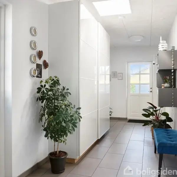 En lys entré med en plante ved siden af en hvid skabsvæg. En bænk står modsat, og der hænger et rundt spejl over hylder dekoreret med vaser.