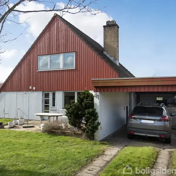 Rødt træhus med skråt tag og skorsten. En bil står parkeret i en carport. Grøn græsplæne og et bord med stole udenfor. Husnummer "24" er synligt.