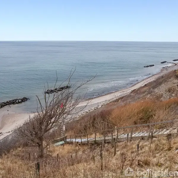 Kyststrækning med grusstier og trappenedgang, der fører til en sandstrand. Havet ligger roligt i baggrunden. Vegetation omgiver stien på en bakke foroven.