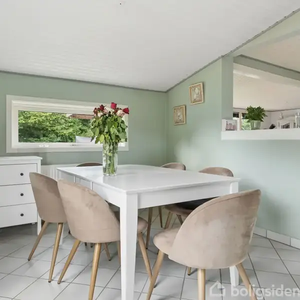 Et hvidt spisebord omgivet af beige stole står i et lyst rum med lysegrønne vægge. På bordet står en vase med røde og hvide blomster. En hvid kommode er placeret nær vinduet.