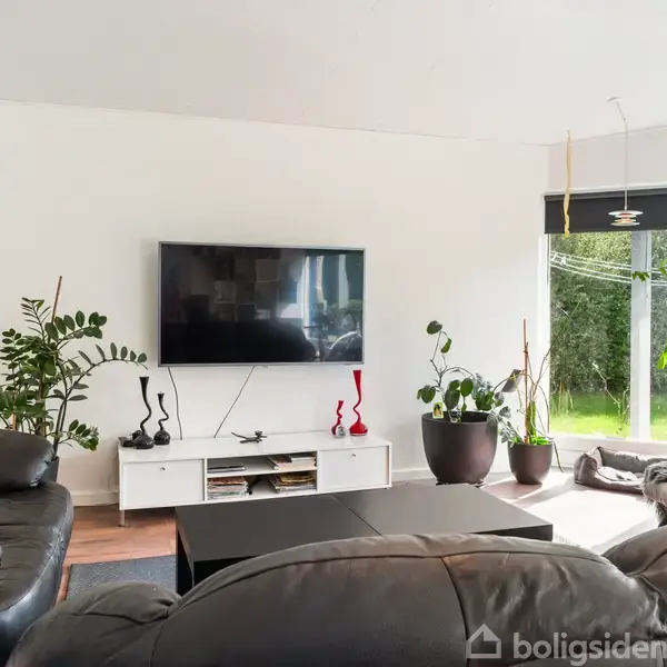 En stue med sorte lædersofaer omkring et sofabord. Et TV hænger på væggen, og der er grønne planter ved det store vindue, der viser en have udenfor.