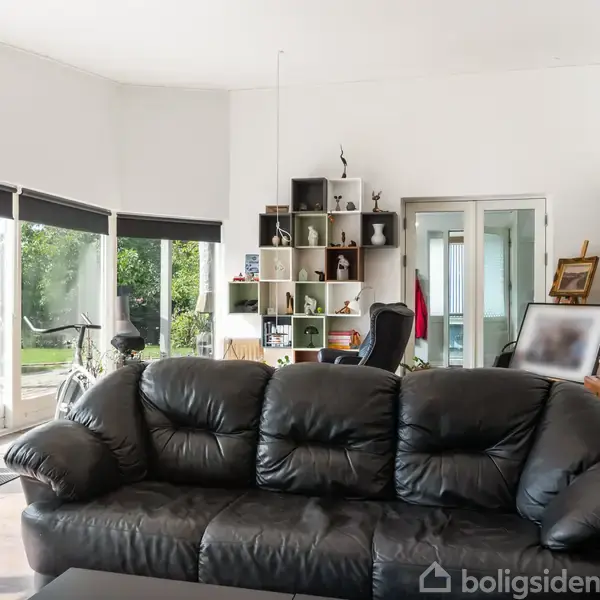Sort lædersofa står foran vinduer med udsigt til grøn have. I baggrunden ses en reol med dekorationer ved siden af en kontorstol og skrivebord.