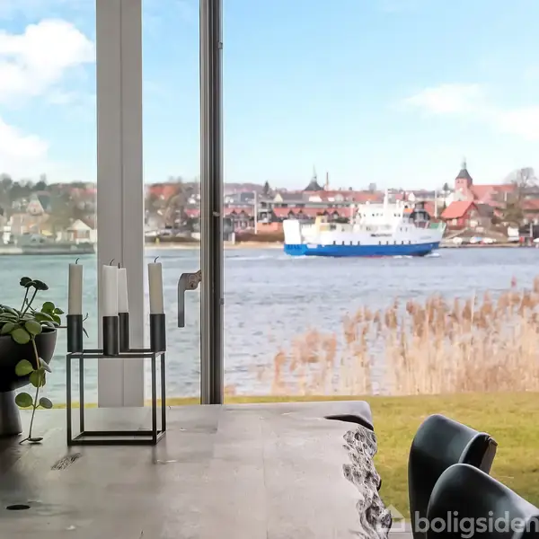 En træbordplade med en plante og lysestager stående. I baggrunden ses en flod med en færge i bevægelse. Bygninger er synlige på den modsatte bred.