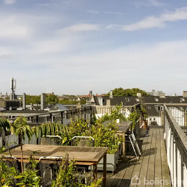 Tagterrasse med borde og stole står tæt ved planter, omgivet af bybygninger i det fjerne under en lys himmel.