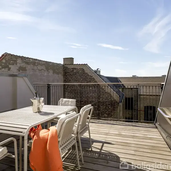 En udendørs tagterrasse indeholder et hvidt bord med stole omkring. En orange genstand ligger på bordet. En håndvask i rustfrit stål er monteret til højre. Bygninger ses i baggrunden.