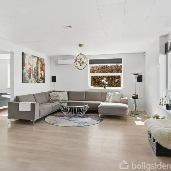 En lys stue med en stor grå hjørnesofa centralt placeret omkring et rundt sofabord. Ved siden af er der store vinduer, der lukker naturligt lys ind. Der hænger kunst på væggene.