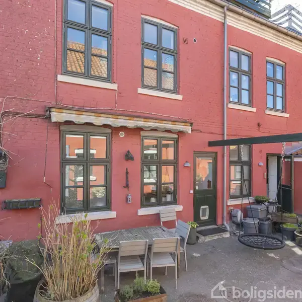 Rødt hus med sorte vinduesrammer har en gårdhave med spisebord, stole og planter i krukker. Omgivelserne inkluderer en overbygning og indgangsparti med flisebelægning og småbede.