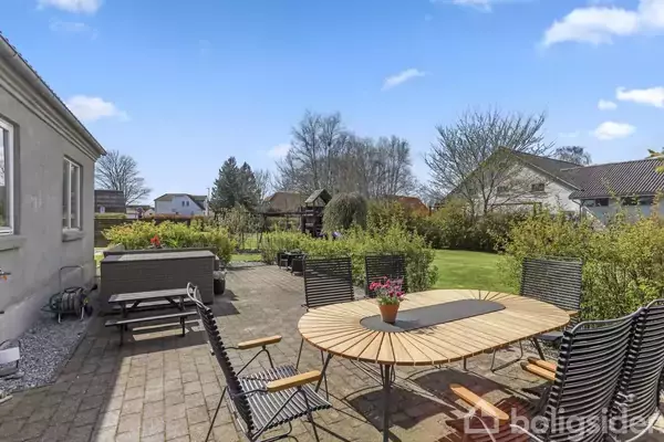 Langbord med stole står på terrasse omgivet af grønne buske og en velplejet græsplæne. I baggrunden ses flere boliger og træer under en blå himmel med spredte skyer.