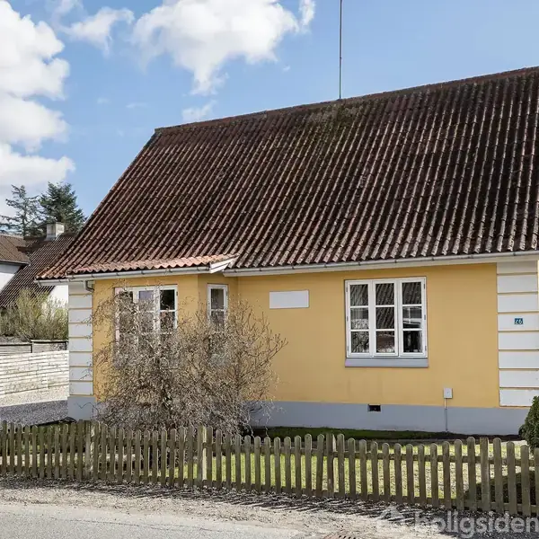 Et gult hus med mørkt tag står bag et hegn og nogle buske. En lille havesektor omkranser huset. Skiltet ved vejen viser "Til salg".