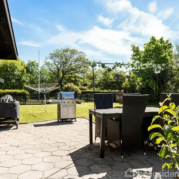 Terrasse med et bord og stole, hvor der står en gasgrill ved siden af. Baggrunden er en grøn have med træer og en tøjstativ under en blå himmel.