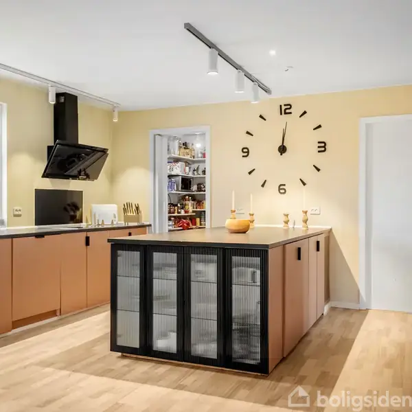 Køkken med brunlige skabe, en central køkkenø med glasdøre, og et stort ur på væggen. Vinduet til venstre giver naturligt lys, og en åben dør viser ind til et spisekammer.