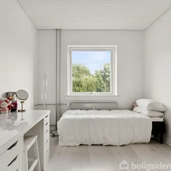 Et enkelt soveværelse med en seng dækket af en hvid dyne. Ved siden af står et skrivebord med en stol og dekorationer. Et vindue giver udsigt til grønne træer udenfor.