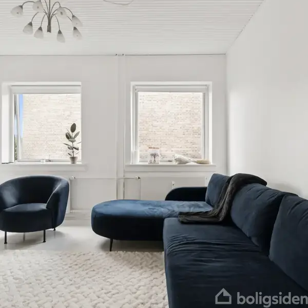 Mørk blå sofa og lænestol står i hvidt rum med et lyst tæppe på gulvet. To vinduer lysner rummet, og der står en plante i vindueskarmen. Lampen hænger i loftet.