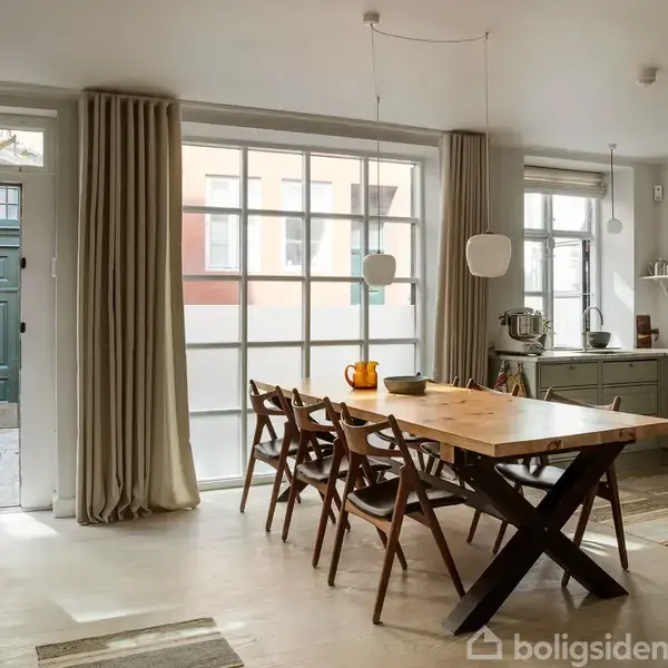Et spisebord med træstole står i et lyst køkken med store vinduer og lyse gardiner. Køkkenet har grønne skabe, åben hylde med porcelæn, og en opsats med frugtskål.