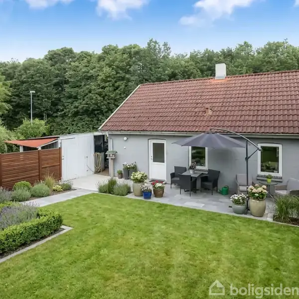 Stue med brændeovn i gang, stabel af brænde ved siden af, spisebord ved vindue, hvide vægge og træloft. Lysindfald fra vinduer skaber en hyggelig atmosfære.