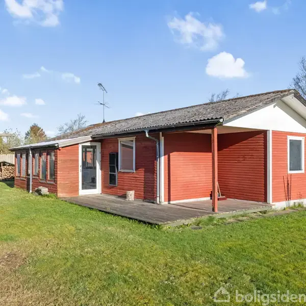Rødt hus står stille på en velplejet græsplæne, omgivet af et hegn og træer under en klar blå himmel.