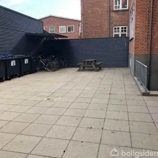 Affaldscontainere står opstillet langs en mur i en gårdhave med fliser. Cykler er parkeret ved siden af. En træg bænk er placeret i hjørnet mod en baggrund af murværk.