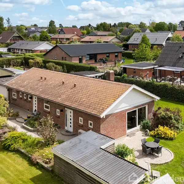 En murstensvilla med tegltag står blandt velplejede haver; en lille terrasse med bord og stole ligger til højre; rækkehuse og grønne træer ses i baggrunden.
