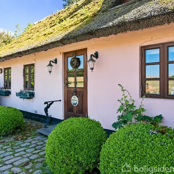 Stråtækt hus med rosa puds, dekoreret med svaler, står langs en brostensbelagt sti omgivet af grønne buske i en solrig have. Text på døren: "Privat" og "25".