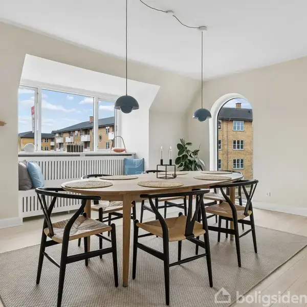 Et spisebord med seks stole står på et lyst tæppe. To pendellamper hænger over bordet. Rummet har lyse vægge, et stort vindue, og en buet døråbning.