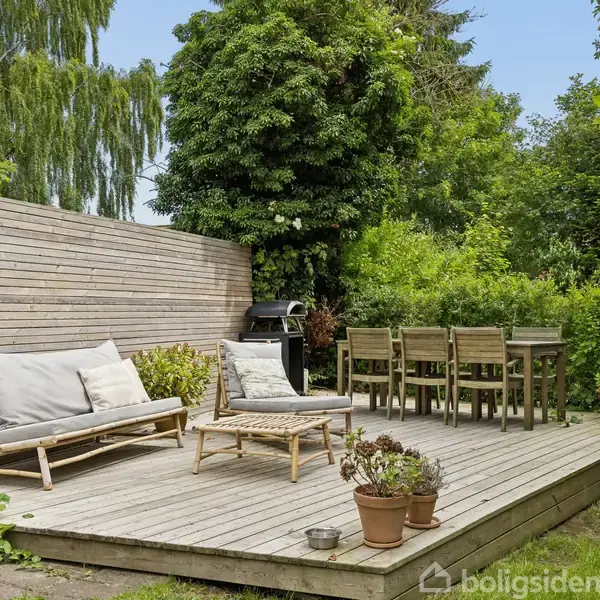 Træterrasse med møbler placeret ved siden af en grill. To liggestole og et spisebord med seks stole står under åben himmel omgivet af tæt grøn vegetation.