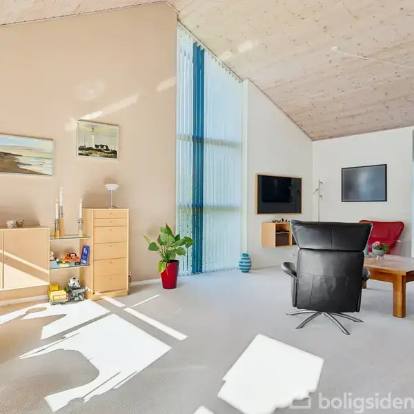 Stue med lyse møbler og kunstgenstande, omfatter reol, planter og lænestol. Store vinduer lader sollys skinne ind, med træbeklædt loft og vægdekoration. Indretningen fremstår moderne og lys.