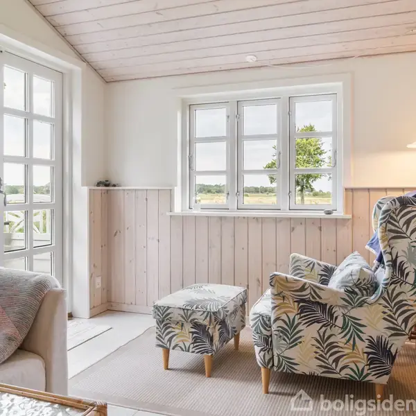 En polstret lænestol med bladmønster står ved et lavt bord i en lys stue. Store vinduer og en glasdør giver udsigt til træer og det åbne landskab udenfor.