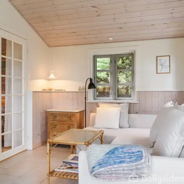 Sofa med puder står i en lys stue ved siden af et vindue. En plaid ligger på sofaen. Bagved er der et lille bord, lampe og en kommode. Velholdt trævæg og træloft skaber en hyggelig atmosfære.