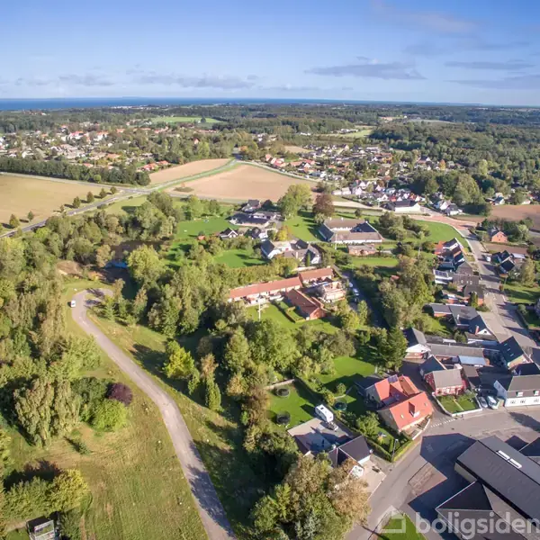 Luftfoto af en lille by med spredte huse omgivet af grønne marker og skove. Veje snor sig gennem byen, som er beliggende i et naturskønt landskab under blå himmel.