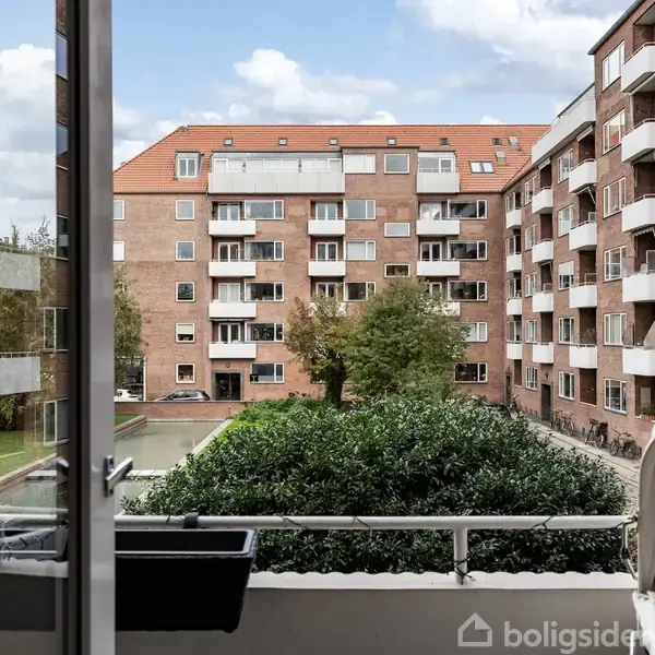 En altan med blomsterkasser kigger ud over en gårdhave med grønne buske og et stort træ. Flankeret af flere etager høje murstensbygninger med balkon. Himlen er delvist overskyet.