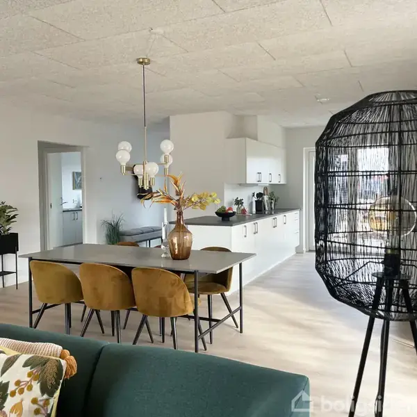 Et spisebord med gule stole står under en moderne lampe i en åben plads. Til venstre er en køkken med hvide skabe og planter. En sort lampeskærm ses i forgrunden.
